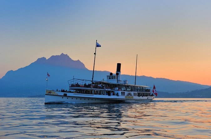 A white paddle steamer on a calm lake at dusk, a jagged mountain behind it