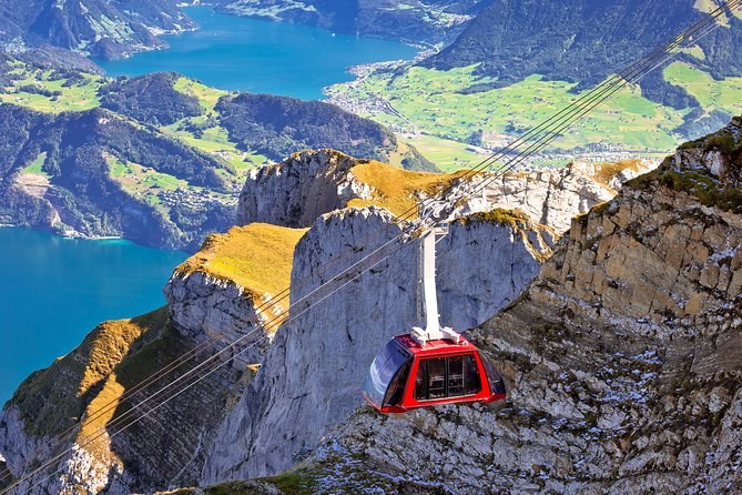 A red cableway cabin crossing above a rocky ridge, with a blue lake and farmland below