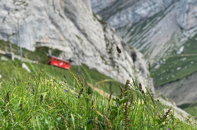 A small red railcar climbing a steep grassy rock face below grey cliffs