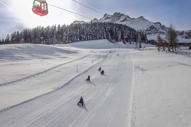 Sledgers on a wide snowy slope, a red gondola cabin passing overhead