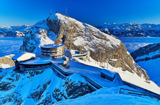 A curved summit hotel and terraces on a snow-covered rock peak under a deep blue sky