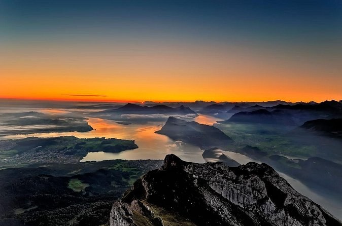 An orange sunset over lakes and hills, seen from a dark rocky summit